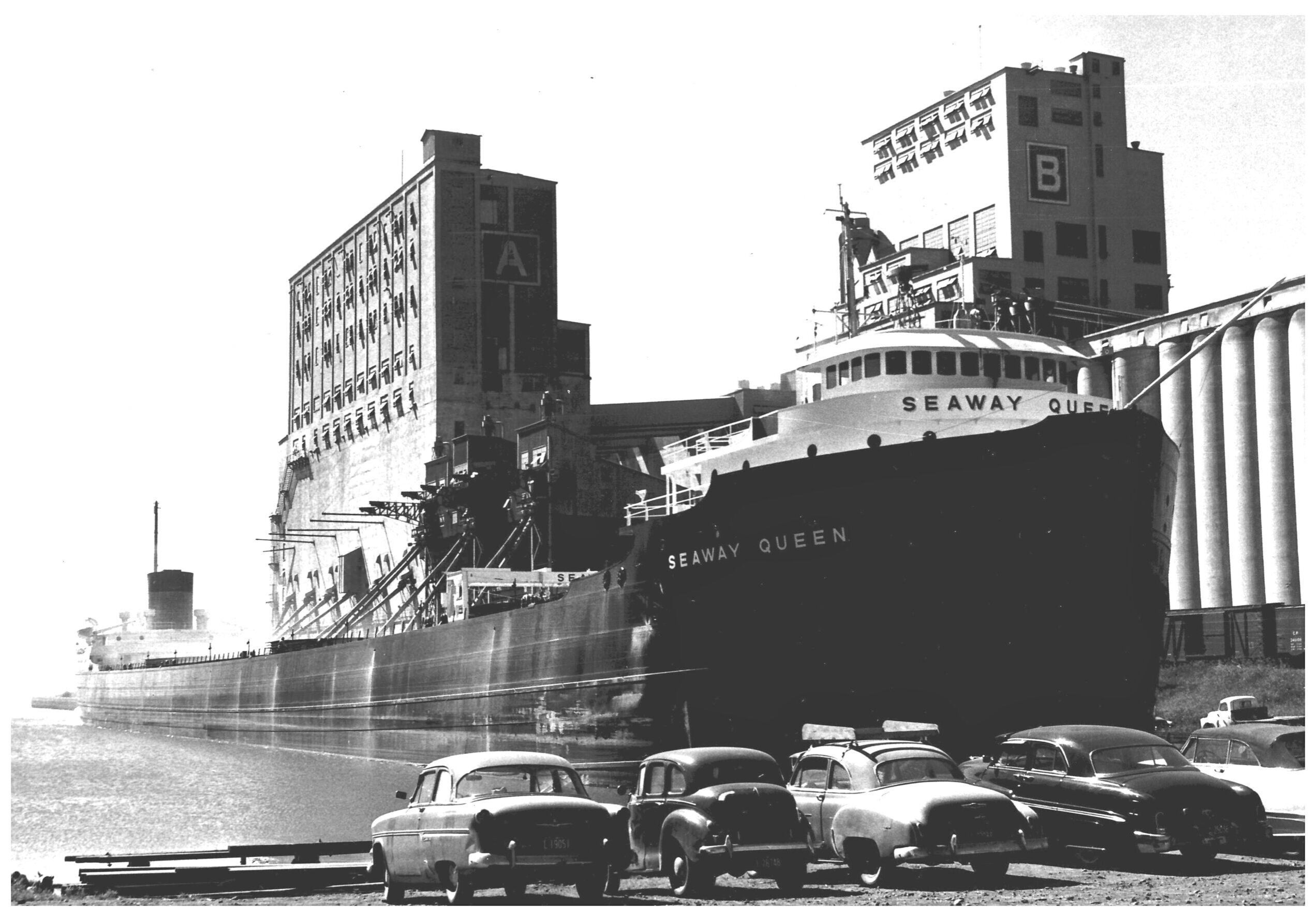 black and white image of cars parked in front of the water with a boat docked in behind, and elevator in the background
