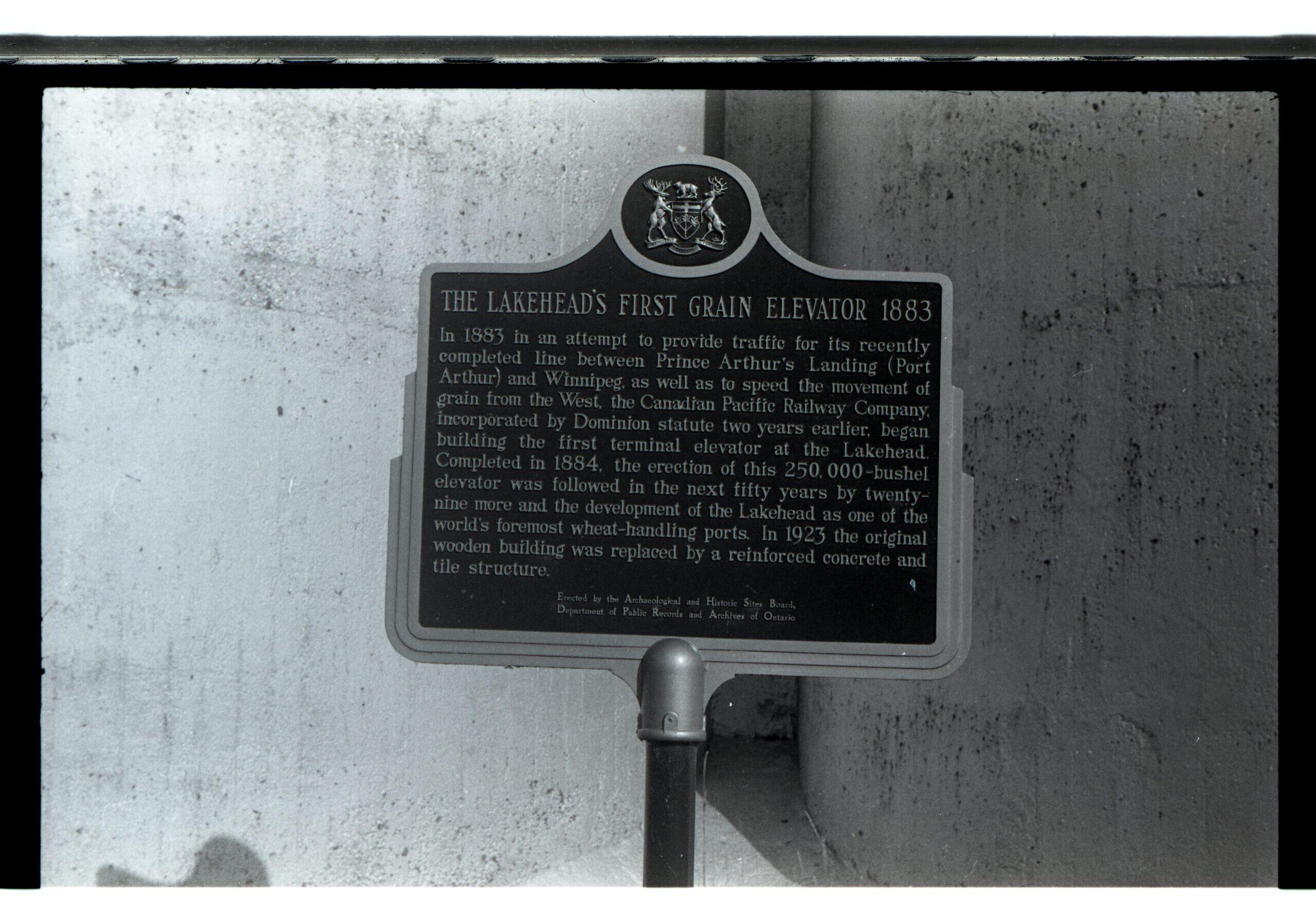 Signage with embelem of Ontario Coat of Arms. Sign reads "THE LAKEHEAD'S FIRST GRAIN ELEVATOR 1883. In 1883 in an attempt to provide traffic for its recently completed line between Prince Arthur's Landing (Port Arthur) and Winnipeg, as well as to speed the movement of grain from the West, the Canadian Pacific Railway Company incorporated by Cominion statue two years earlier, began building the first terminal elevator at the Lakehead. Completed in 1884, the erection of this 250,000-bushel elevator was followed in the next fifty years by twenty-nine more and the development of the Lakehead as one of the world's foremost wheat-habdling ports. In 1923 the original wooden building was replaced by a reinforced concrete and tile structure"
